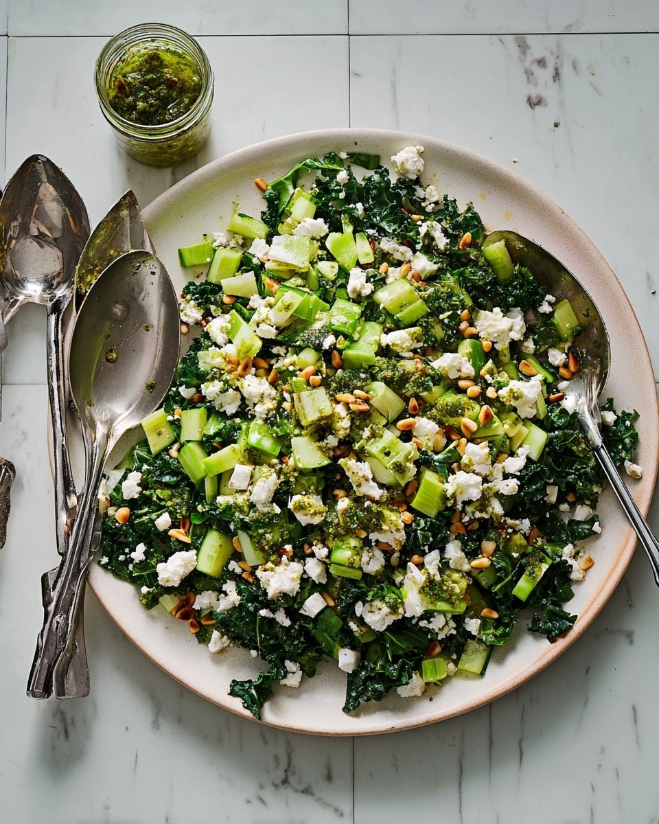 A large white plate holds a fresh salad made of several layers: a base of dark green kale leaves, mixed with light green chopped celery and cucumber pieces, topped with white crumbled cheese scattered unevenly, and sprinkled with small light brown pine nuts. There are also small bright green onion slices and spots of green pesto sauce spread on top. The plate rests on a white marbled surface alongside two shiny silver salad serving spoons, and a small jar with more green pesto is visible above the plate. photo taken with an iphone --ar 4:5 --v 7