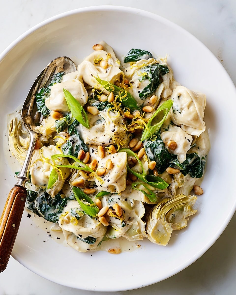 The dish shows a white round plate filled with creamy tortellini pasta coated in a smooth white sauce, mixed with dark green leafy spinach and pale yellow artichoke pieces, creating a layered effect. On top, golden toasted pine nuts are scattered along with bright green fresh scallion slices and a light dusting of black pepper and lemon zest, adding texture and color contrast. A silver fork with a wooden handle rests inside the plate at the left side, all placed on a white marbled surface. photo taken with an iphone --ar 4:5 --v 7
