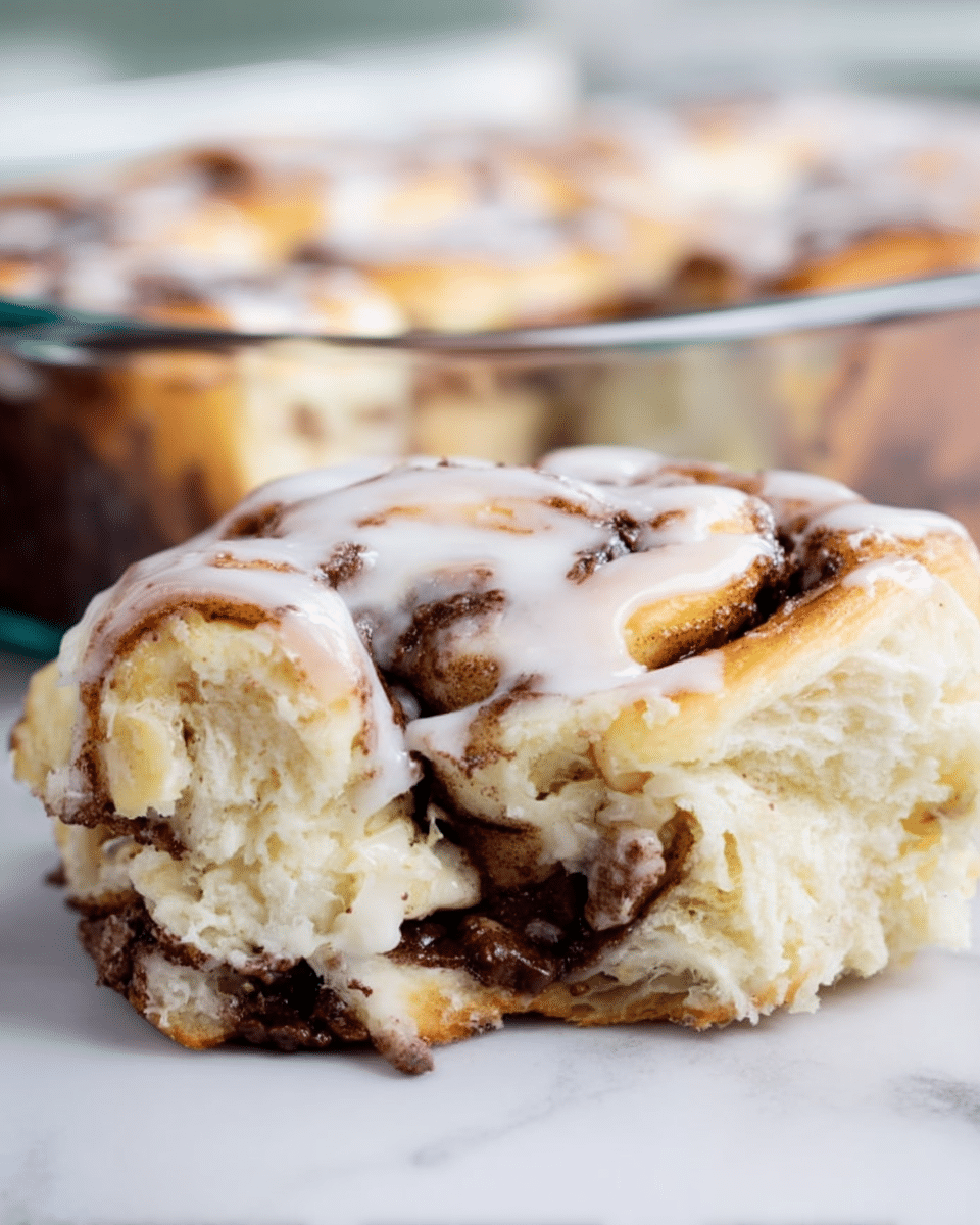 A close-up of a cinnamon roll with two thick layers of soft, light golden dough twisted around a dark, gooey chocolate filling. The top is drizzled with a smooth, white glaze that adds a glossy texture. The roll shows a moist interior with visible swirls of melted chocolate embedded in the fluffy dough. The background features a clear glass baking dish with more cinnamon rolls on a white marbled surface. photo taken with an iphone --ar 4:5 --v 7