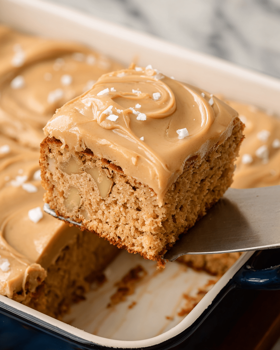 A thick square cake piece is held above a white baking dish by a silver spatula, showing one layer of light brown, soft cake with small chunks inside and a thick top layer of smooth, creamy caramel-colored frosting with a few flakes of sea salt sprinkled on top. The baking dish holds the rest of the cake with the same one-layer cake and frosting, sitting on a white marbled surface. Photo taken with an iphone --ar 4:5 --v 7