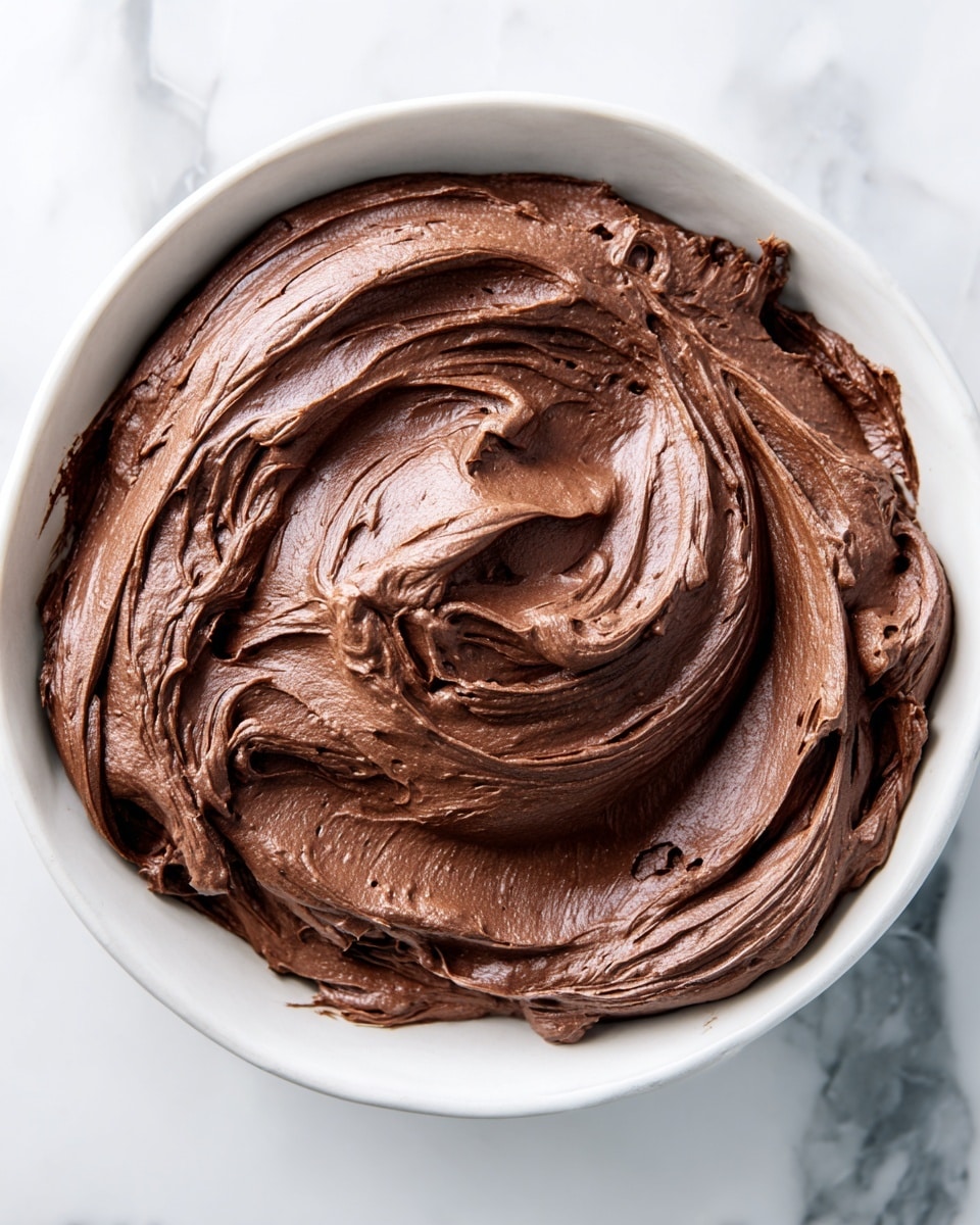 A thick, smooth swirl of rich, dark brown chocolate frosting fills a white bowl, with creamy, soft peaks and waves creating a textured surface that looks glossy and airy. The frosting is piled high with visible spatula marks that form natural curves and folds, giving a sense of depth and lightness. The bowl sits on a white marbled surface that adds a clean and bright contrast to the deep chocolate color. photo taken with an iphone --ar 4:5 --v 7