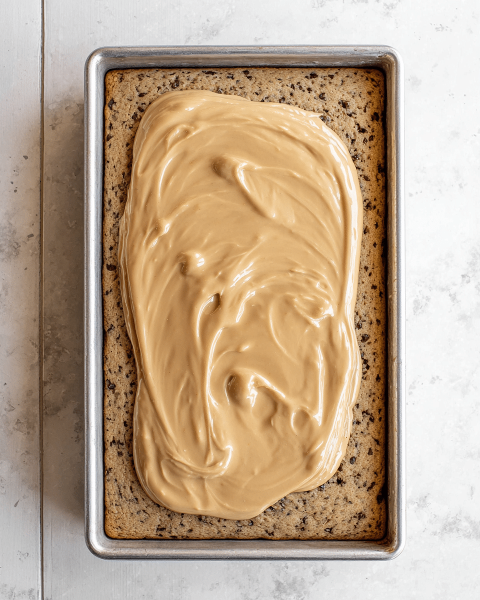 A single square piece of cake is shown on a white plate with a black patterned rim, resting on a white marbled surface. The cake has two visible layers: the bottom layer is light brown and moist with small chunks, and the top layer is a smooth light tan frosting spread evenly across. Crushed nuts, in medium to small pieces with a rough texture and a mix of light and dark brown colors, are sprinkled over the frosting. A silver fork rests on the plate, holding a small bite of the same cake. Photo taken with an iphone --ar 4:5 --v 7