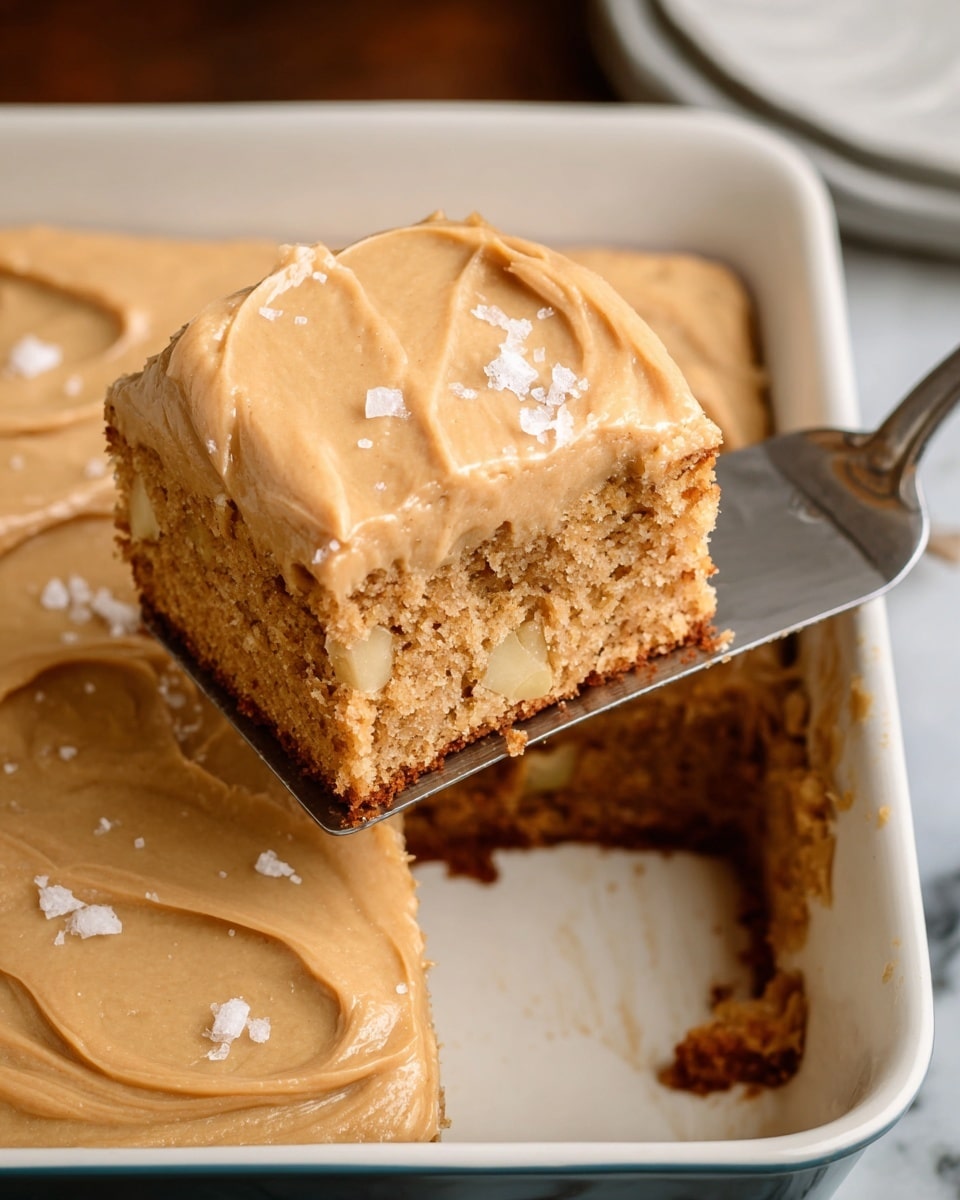 A close-up of a single square piece of light brown cake with small chunks inside being lifted by a metal spatula from a larger cake in a white baking dish with dark blue edges. The cake has two visible layers: a thick, spongy base with a slightly rough texture and a smooth, creamy caramel-colored frosting layer on top. The frosting is decorated with uneven swirls and sprinkled with small white flakes. The background is a white marbled surface. Photo taken with an iphone --ar 4:5 --v 7