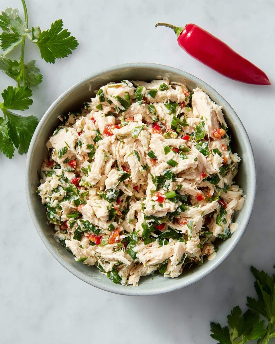 A close-up view of a single-layer dish in a round white bowl filled with shredded white chicken mixed with finely chopped green herbs and small bits of red chili peppers, all stirred together to make a textured and colorful chicken salad. The bowl is placed on a white marbled surface with a bright red chili pepper and fresh green parsley leaves nearby. The image captures a fresh and simple meal with light colors and a natural look. photo taken with an iphone --ar 4:5 --v 7